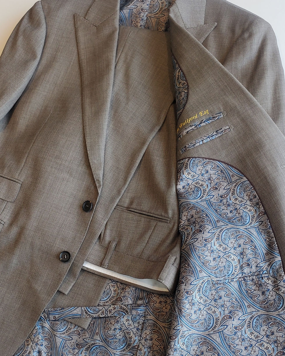 A soft brown original suit, designed by Jo. - with French Interior and Paisley Lining, piped in a chocolate satin, with a gold embroidered inscription and a visible vent strap.