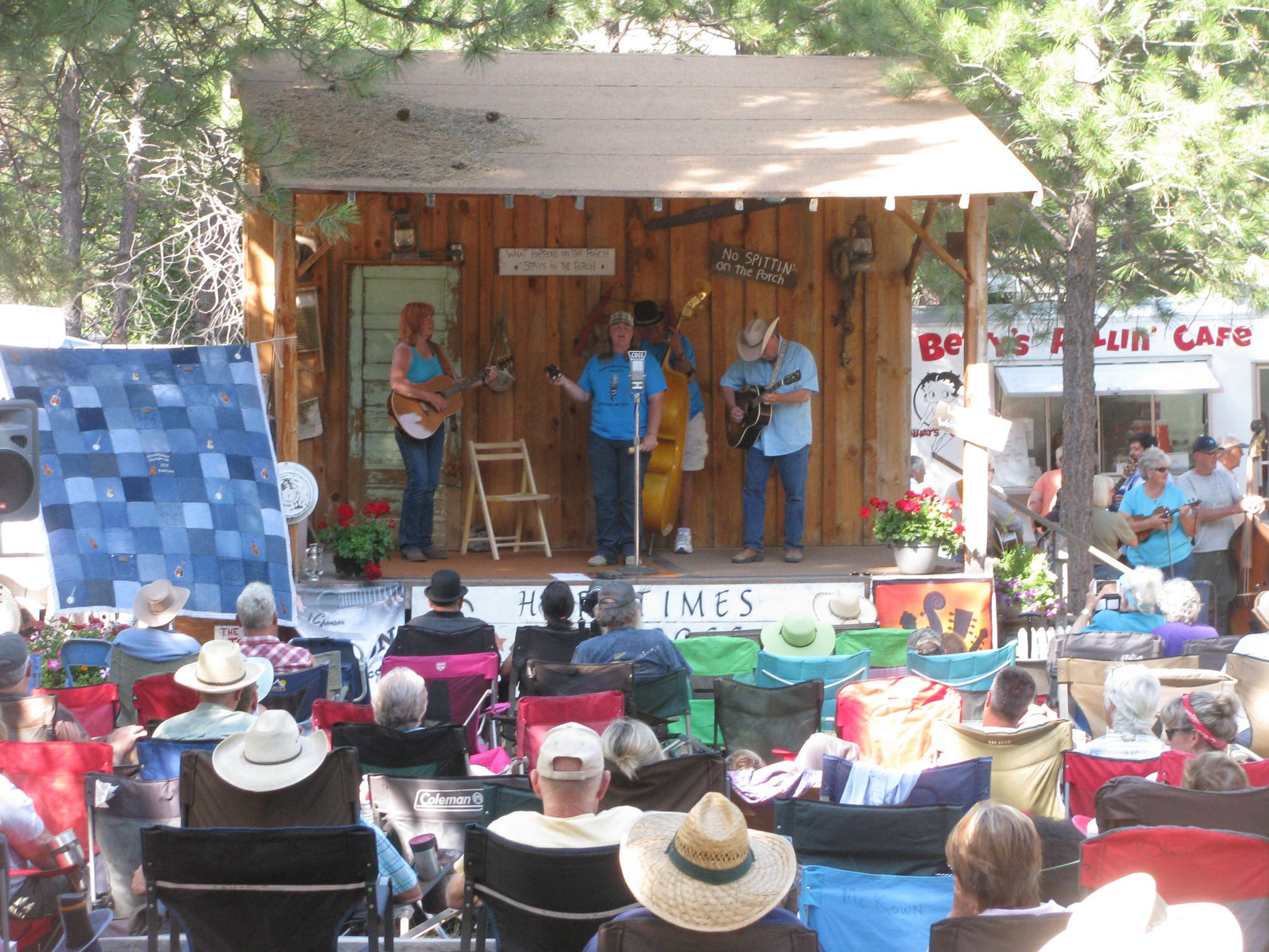 Bluegrass musicians performing on a small outdoor wooden stage for a seated audience.