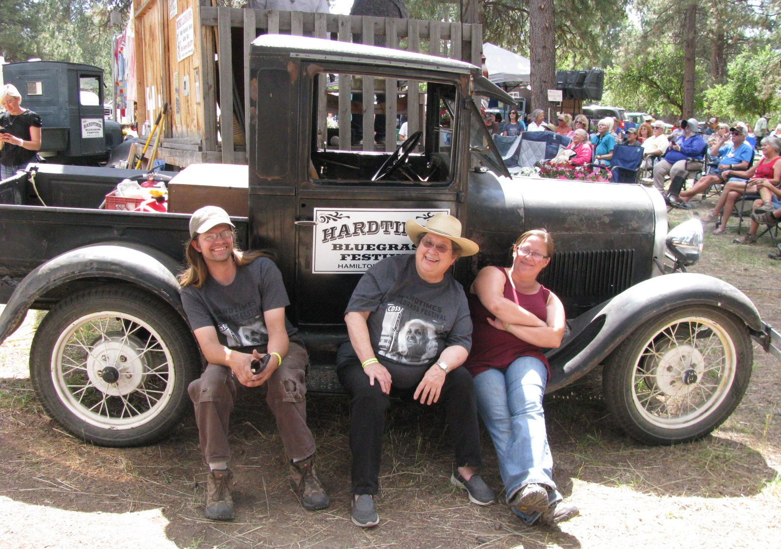 Three people sitting in front of a vintage truck at an outdoor festival, with seated attendees behind them