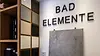 Badwissen von den Profis: Das Logo von BAD ELEMENTE an einer Ausstellungswand mit verschiedenen Materialmustern für Fliesen und Oberflächen – fundierte Tipps und Trends aus über 25 Jahren Erfahrung in der Badsanierung.