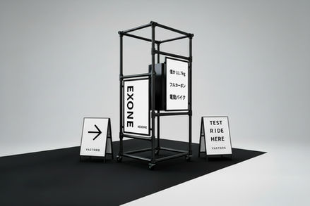 単管パイプで組まれた立体フレームの展示ディスプレイ。中央に「EXONE」のポスターと「僅か11.7kg フルカーボン 電動バイク」のパネルを配置し、周囲には「TEST RIDE HERE」のサインスタンドが置かれている、ミニマルな展示構成。