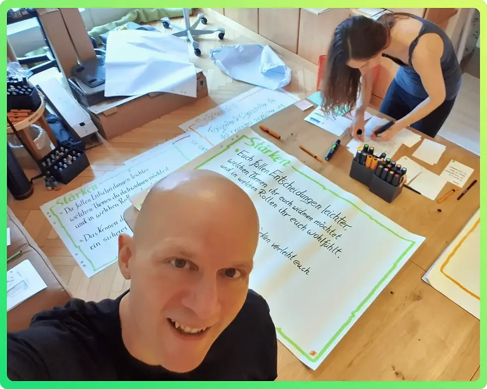 Ein Selfie von Sandor (Vordergrund, lächelnd), während Conny im Hintergrund am Boden und auf einem Tisch an großen Postern arbeitet. Auf den Postern ist das Wort "Stärken" und handgeschriebene Business-Konzepte zu sehen.