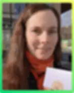 Smiling woman with long hair, wearing an orange scarf, holds a white paper with text "Moment F...". Reflections in glass behind her.