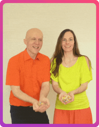 Conny, eine lachende Frau mit langen braunen Haaren in einem neongelben Oberteil und einem orangefarbenen Rock, und Sandor, ein lachender, glatzköpfiger Mann in einem orangefarbenen Hemd, spielen mit buntem Konfetti.