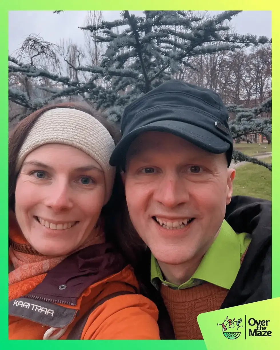 Conny, eine lächelnde Frau mit braunen Haaren und einem Stirnband, und Sandor, ein lächelnder, glatzköpfiger Mann mit schwarzer Kappe, posieren für ein Selfie im Freien vor Bäumen.