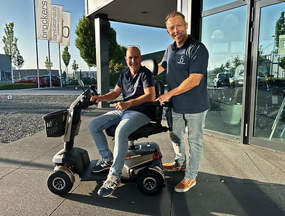 Ein Mitarbeiter sitzt auf einem E-Mobil, während der zweite Mitabeiter dahinter steht. Beide schauen freundlich zur Kamera.