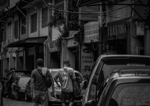 A moody, black-and-white street photograph showing the backs of two men walking down a historic, narrow alleyway lined with parked cars, old shophouses, and vintage storefront signage.
