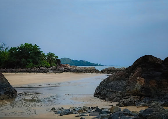Quiet tropical shoreline framed by dark rocks, with wet sand, calm water and a distant forested headland beneath a muted blue-grey sky.