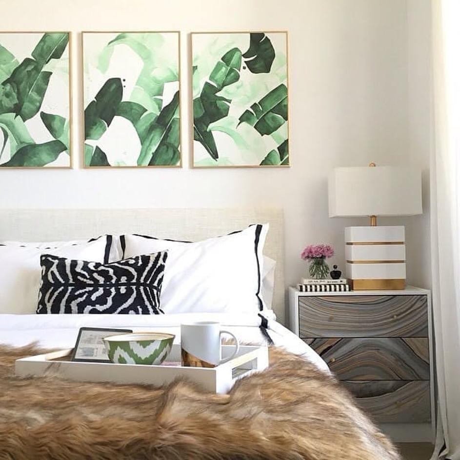 A bedroom with a cozy bed and three frames above it with green palm leaves wallpaper.