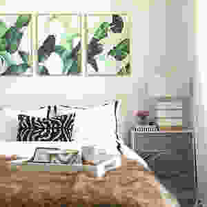 A bedroom with a cozy bed and three frames above it with green palm leaves wallpaper.