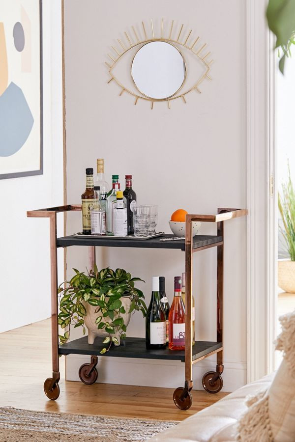 A rolling wooden bar cart from Urban Outfitters with alcohol and plants under a small gold mirror shaped as an eye hanging on the wall.