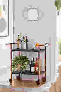 A rolling wooden bar cart from Urban Outfitters with alcohol and plants under a small gold mirror shaped as an eye hanging on the wall.