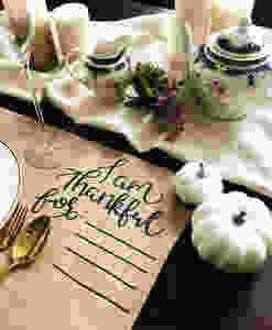 A kraft placemat that reads, I am thankful for, with empty lines next to white pumpkins, gold utensils, and tea.