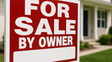For Sale By Owner" sign staked in front of a suburban house with green lawn and blue sky.
