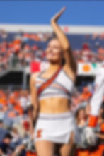 Cheerleader in a white and orange outfit waves and smiles during a sunny outdoor event. Crowd and stadium in the blurred background.