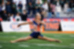 A baton twirler in a navy blue and orange outfit performs a split on a football field with a crowd in the background.