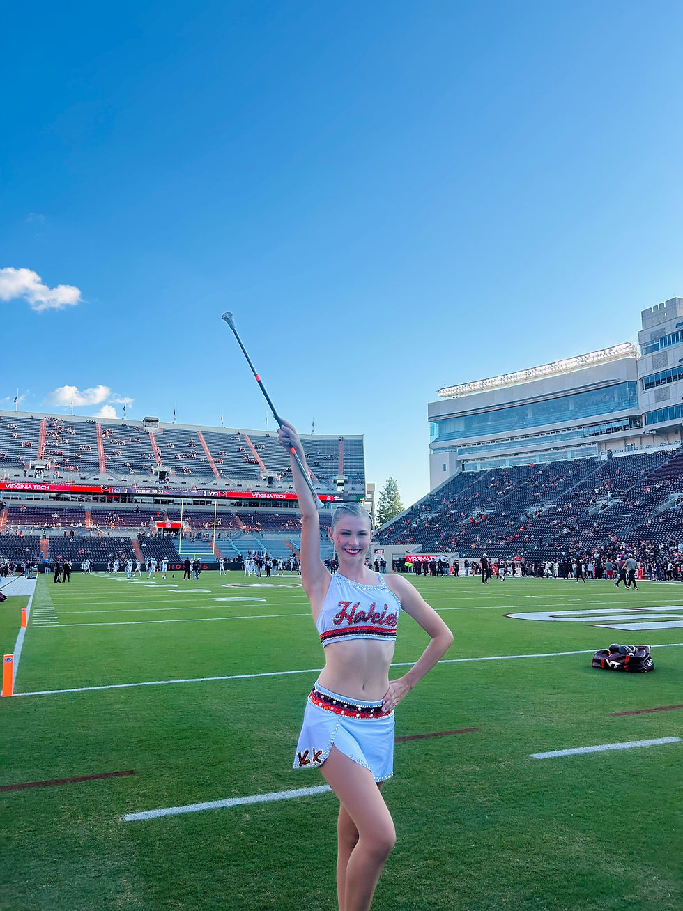 Cheerleader in a white uniform with "Hokies" text poses on a football field, holding a baton aloft. Stadium and fans in background.