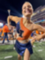 Cheerleader in orange-blue outfit poses with baton, smiling at a stadium with crowd and bright lights. "I" logo visible on costume.