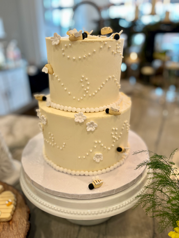 Two-tier white baby shower cake featuring bee details and white floral decorations.