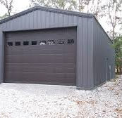 Dark gray metal garage building with brown door and gravel driveway.