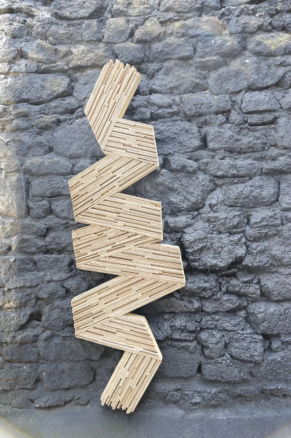 Sculpture en carton ondulé en zigzag contre un mur de pierres. Galerie Mourier
