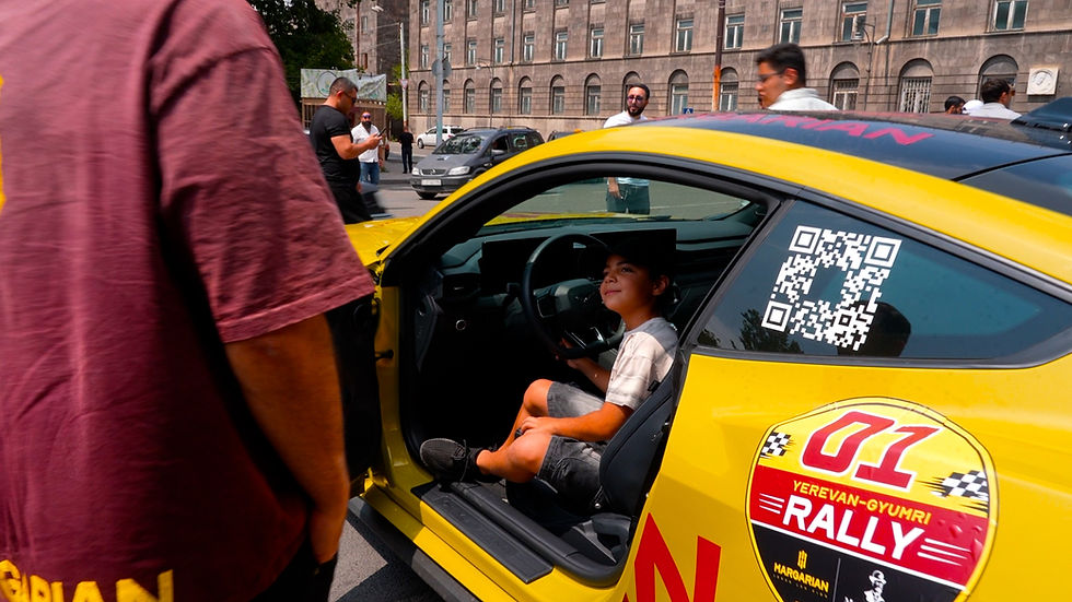 Boy sits in a yellow rally car with open door and QR code, at a Yerevan-Gyumri Rally street gathering with onlookers.