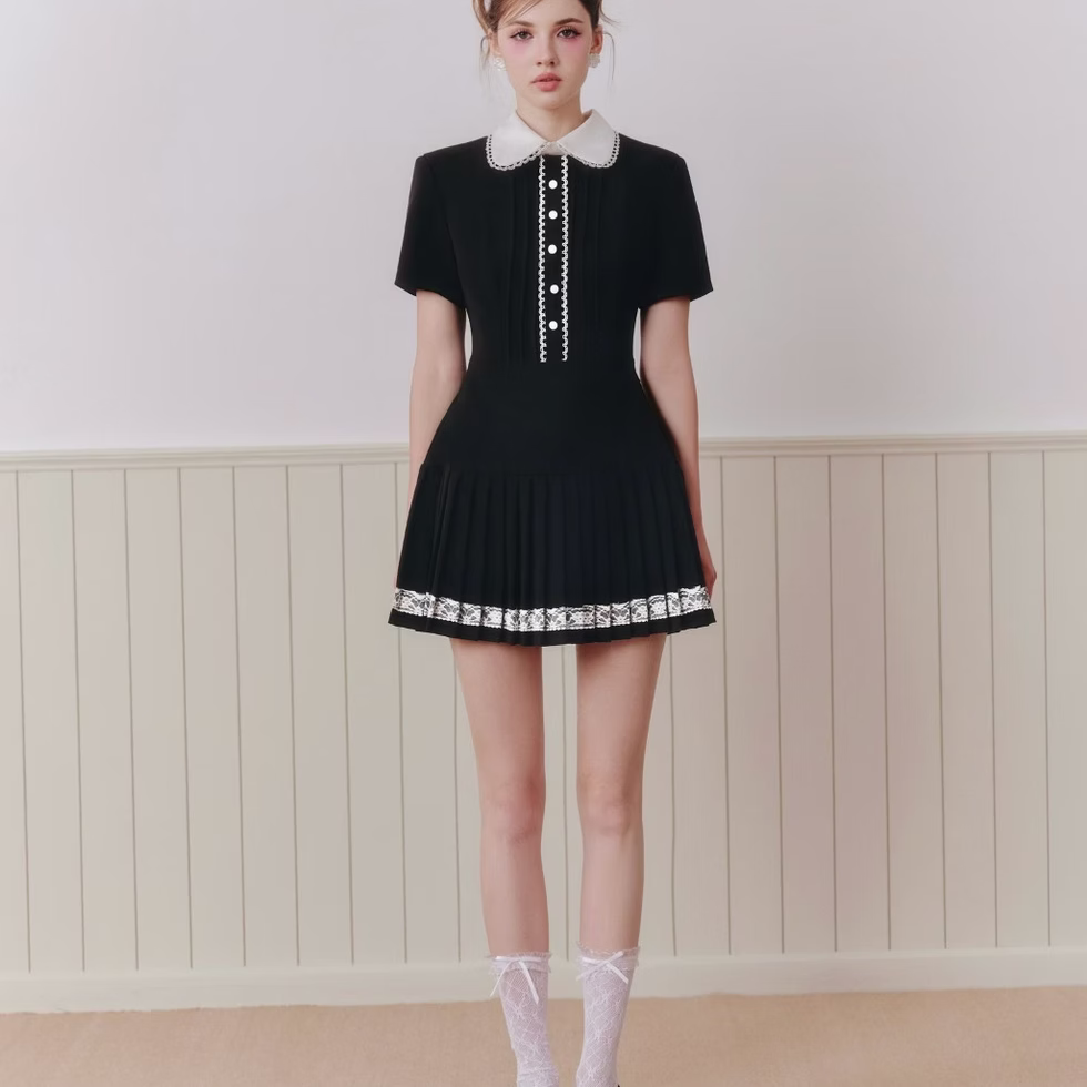 Model wearing black dress with white lace collar, short sleeves, pleated skirt with lace trim, posing in minimal cream room.