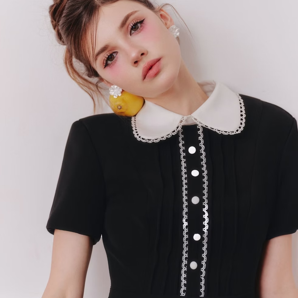 Model wearing black dress with white lace collar, short sleeves, pleated skirt with lace trim, posing in minimal cream room.