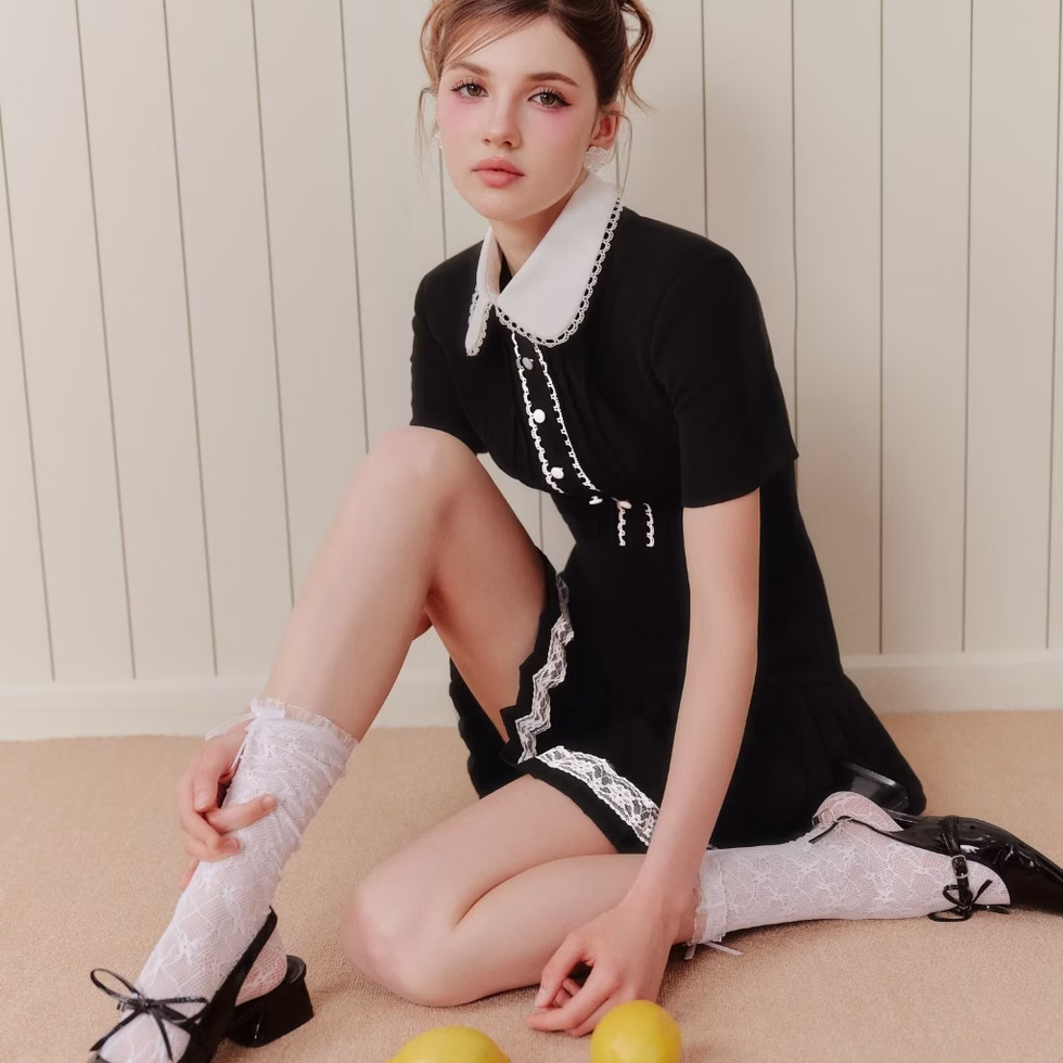 Model wearing black dress with white lace collar, short sleeves, pleated skirt with lace trim, posing in minimal cream room.