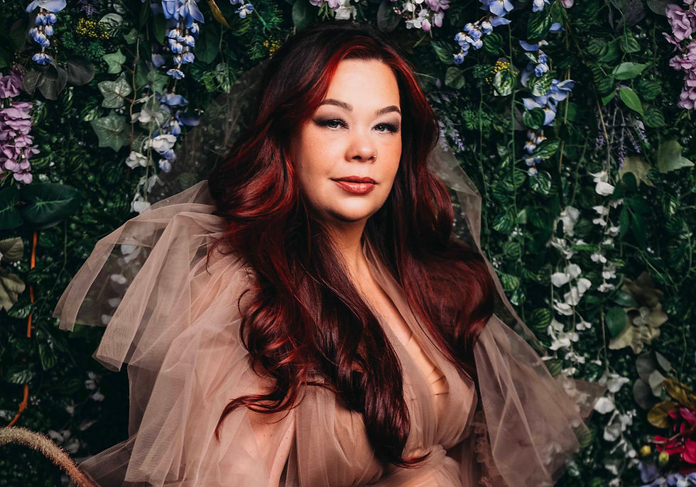 Portrait of a woman with long auburn hair in a sheer beige dress, posed against a lush green and purple floral wall