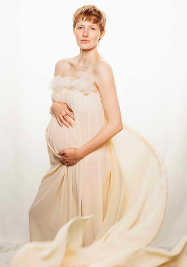 Pregnant woman holding belly against white glowing background as cream fabric swirls around her.