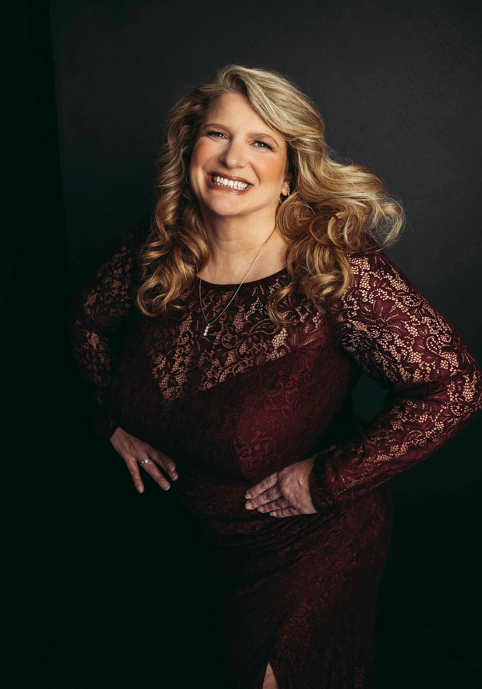 Smiling mid aged woman in a burgundy lace dress poses with hands on hips against a dark background, conveying confidence and elegance.