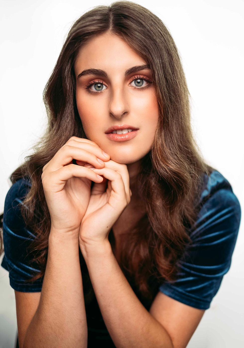 Portrait of a young woman with long brown hair and blue eyes, hands clasped under her chin, in a blue velvet top against white.