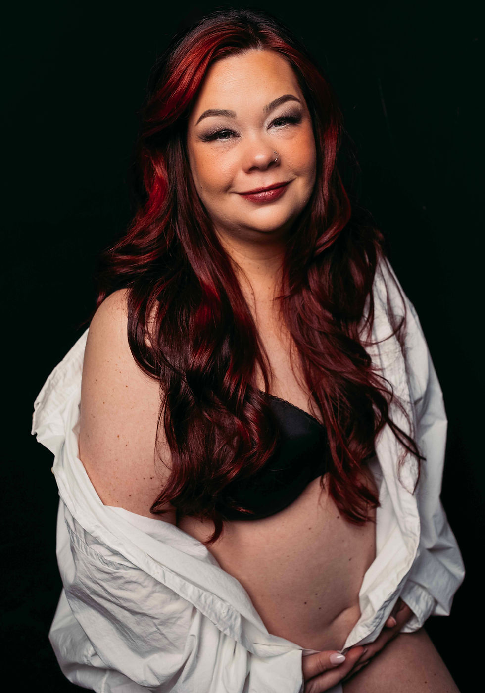 Portrait of a woman with red hair in a white shirt and black top, smiling softly against a dark background.