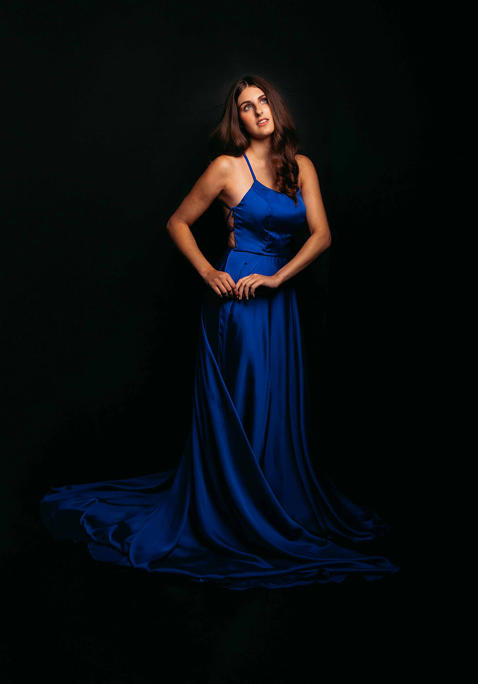 Young woman in a flowing royal blue gown poses against a black studio backdrop, looking upward thoughtfully.