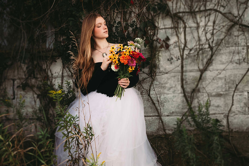 Young woman in black top and white tulle skirt holds a colorful bouquet beside a vine-covered stone wall, eyes closed.