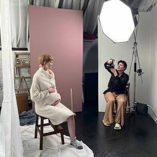 Pregnant woman sits on stool holding her belly as photographer in black shirt photographs her.