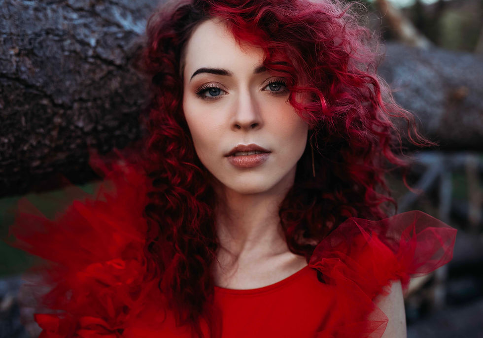 Woman with curly red hair and a red dress stands against a dark textured background, looking intently at the camera.