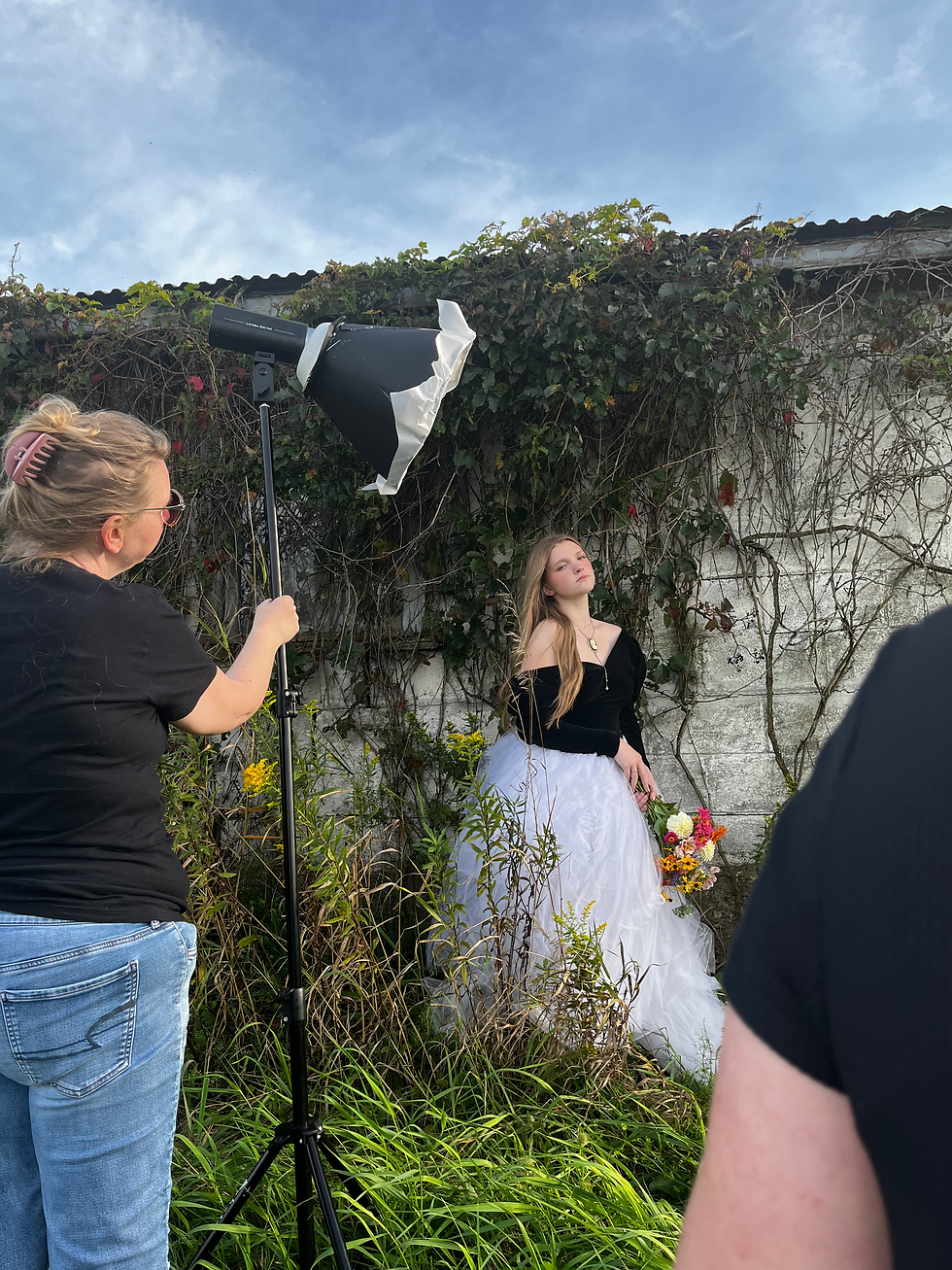 Outdoor photoshoot: a woman adjusts a studio light while a young woman in a black top and white tulle skirt poses with flowers by a vine-covered wall.