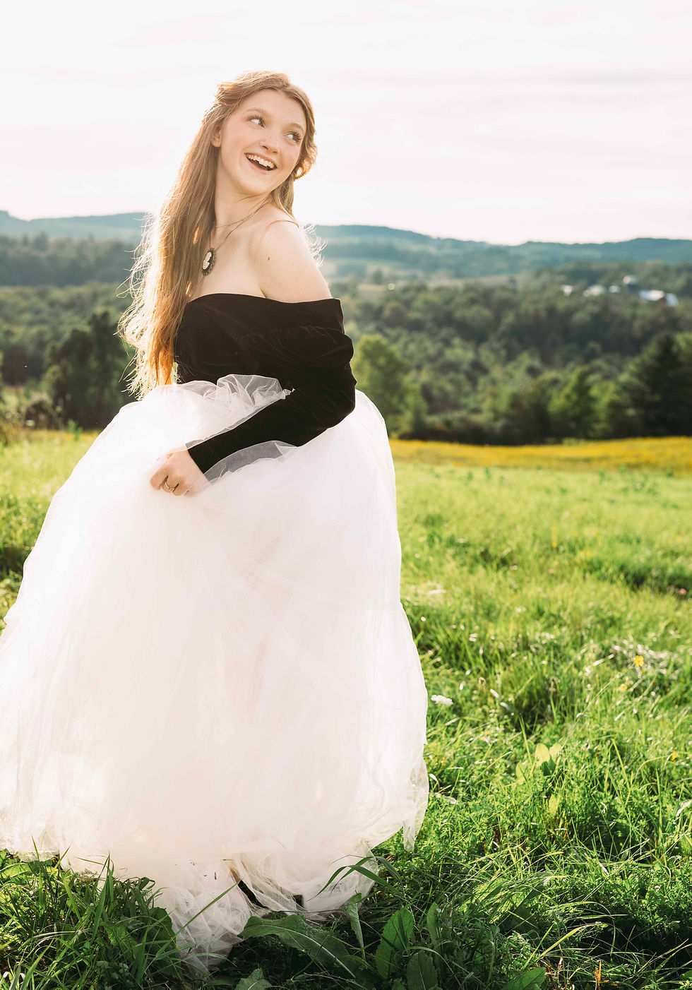 Smiling young woman in a black off-shoulder top and white tulle skirt stands in a sunny meadow with hills behind her.