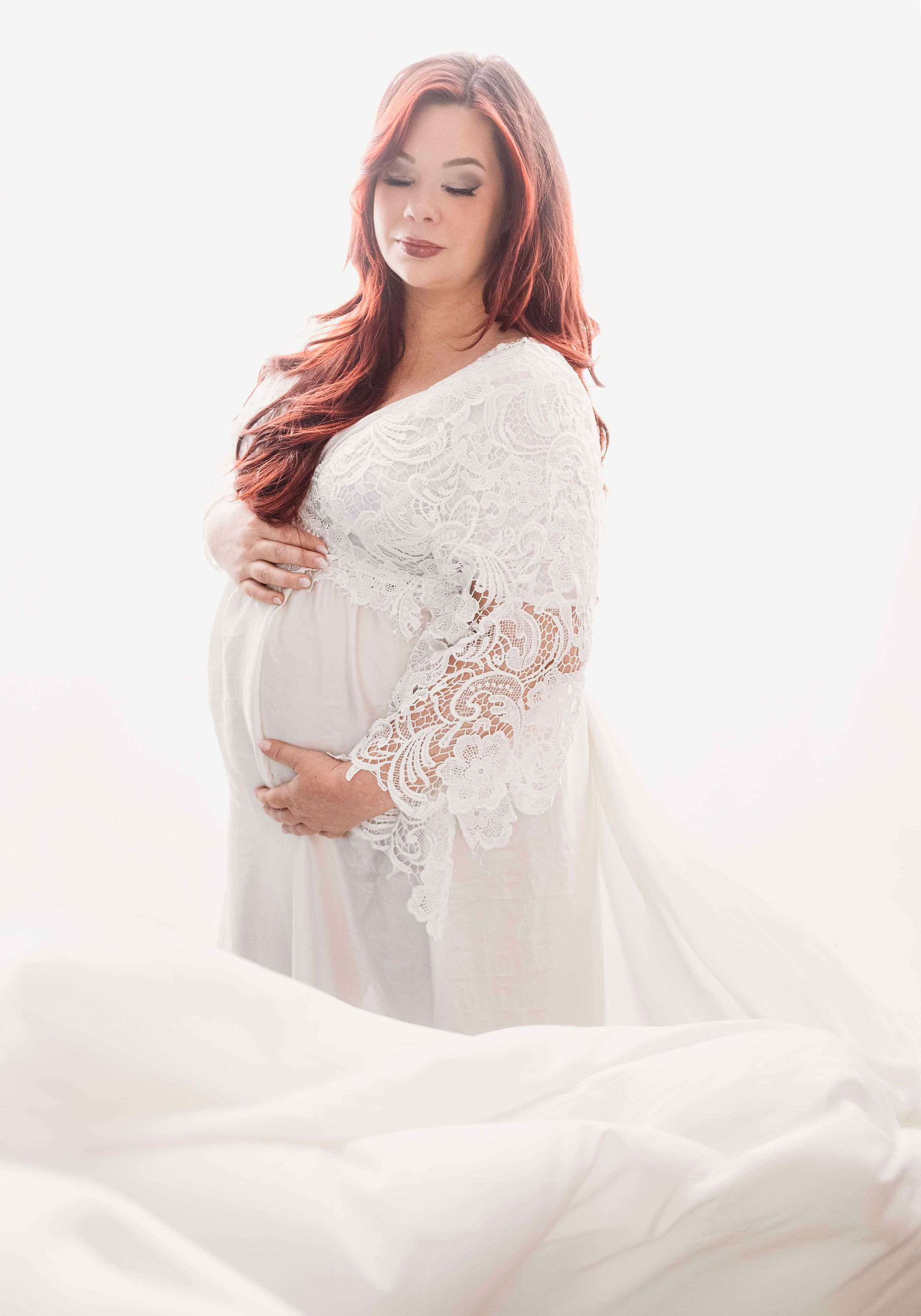 Maternity picture of woman as she holds her belly and looks down. She's surrounded by white lights and she wears a lace top.