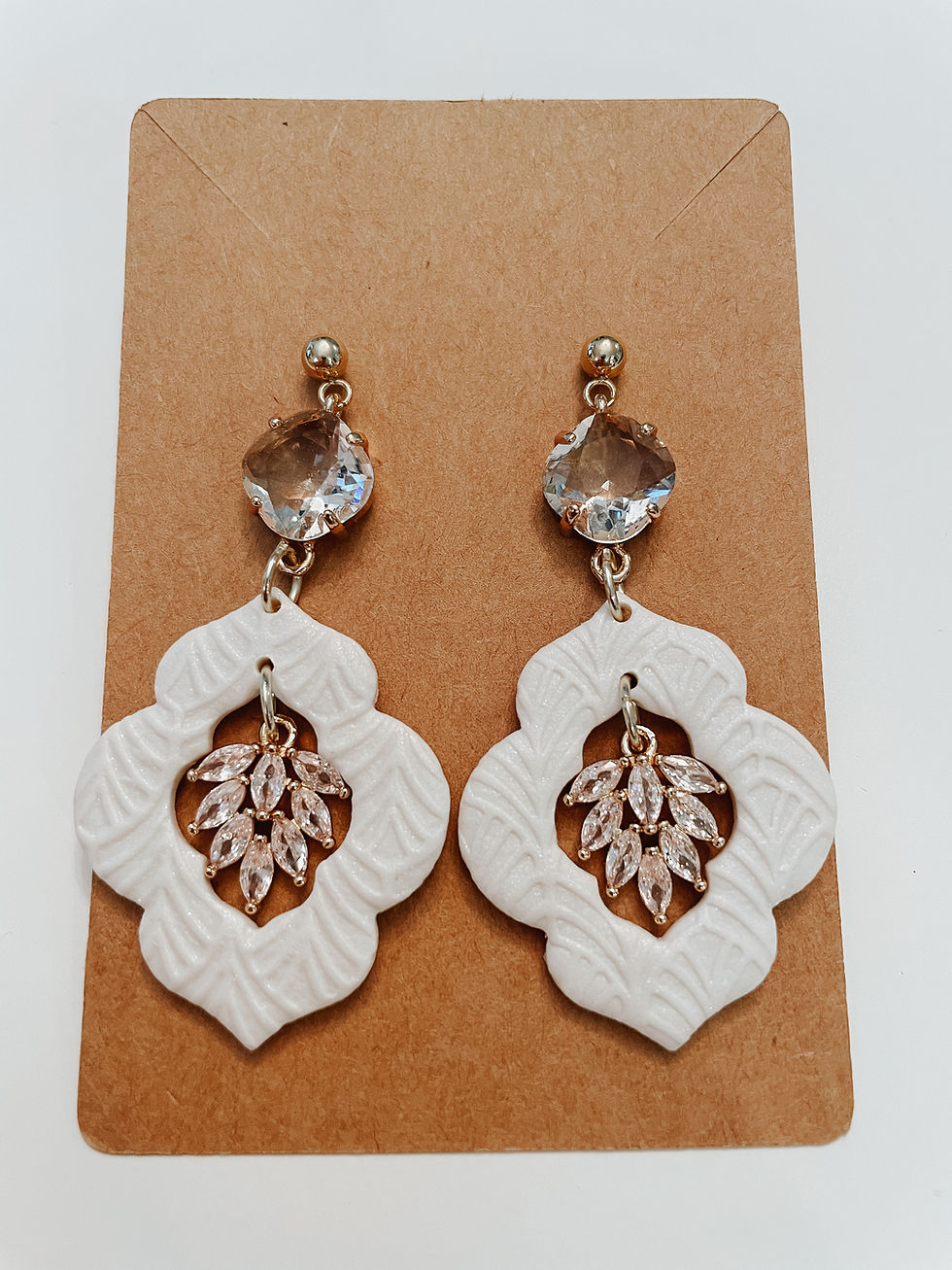 White earrings with crystals and ornate design on a tan background.