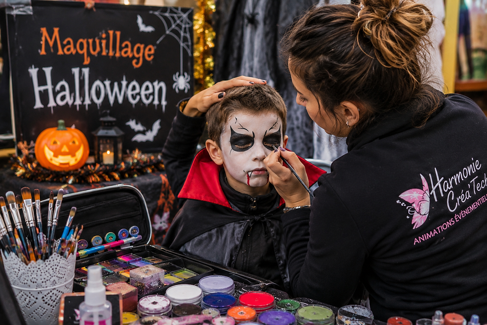 Maquilleuse artistique réalisant un maquillage d'Halloween sur un enfant déguisé en vampire, devant un stand décoré aux couleurs d'Halloween.