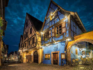 Maison à colombages colorée dans une rue pavée d’un village alsacien de nuit, illuminée par des lumières chaleureuses, avec une terrasse de café sous un ciel bleu profond.