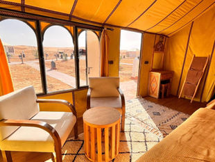 Bright yellow glamping tent interior with armchairs and a round table. Desert scenery visible outside through large windows. Warm and inviting.