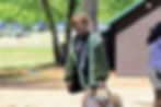 At Camp Waukeela a person in green sweater walking outside near a cabin, carrying a bag. Sunny day, trees and a fence in the background. Casual mood.