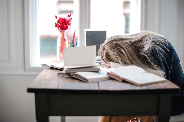 A woman face down on a desk, lying on top of a pile of books. There is a a vase with red flowers in the background.