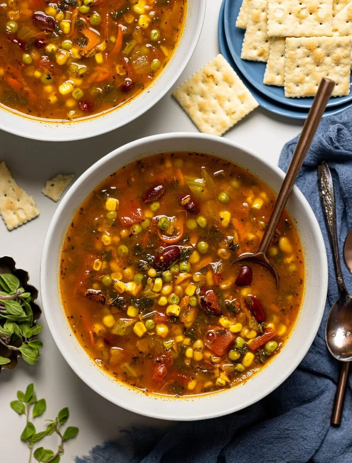 Two bowls of Loaded Vegan Veggie Soup on a white surface with a blue napkin and salt water crackers