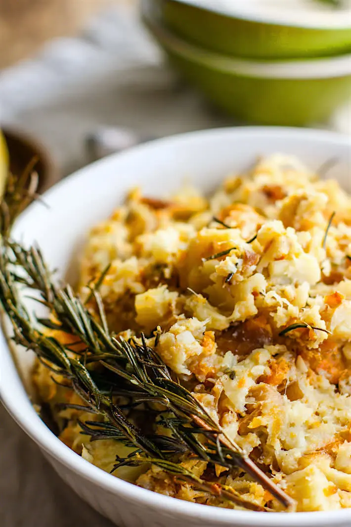 A white bowl with CrockPot Carrot Parsnip Mash and a sprig of fresh rosemary on top. in the background are a stack of green bowls and a white table cloth.