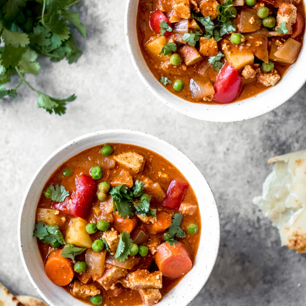 Two white bowls with Vegan Slow Cooker Tofu Tikka Masala on a stone background with fresh parsley in the top right corner